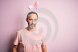Middle age hoary man wearing bunny ears standing over isolated pink background skeptic and nervous, frowning upset because of