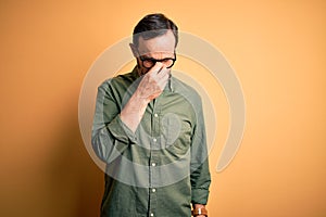 Middle age hoary man wearing casual green shirt and glasses over isolated yellow background tired rubbing nose and eyes feeling