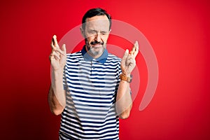 Middle age hoary man wearing casual striped polo standing over isolated red background gesturing finger crossed smiling with hope