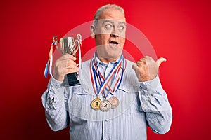 Middle age hoary successful man wearing medals holding trophy over isolated red background pointing and showing with thumb up to