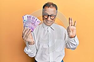 Middle age indian man holding 100 philippine peso banknotes doing ok sign with fingers, smiling friendly gesturing excellent