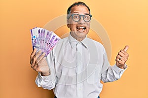 Middle age indian man holding 100 philippine peso banknotes pointing thumb up to the side smiling happy with open mouth