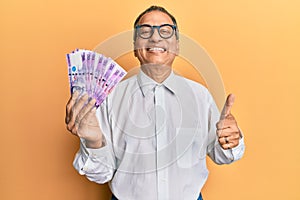 Middle age indian man holding 100 philippine peso banknotes smiling happy and positive, thumb up doing excellent and approval sign