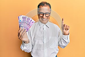 Middle age indian man holding 100 philippine peso banknotes smiling with an idea or question pointing finger with happy face,