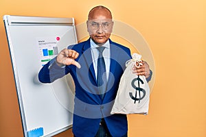 Middle age latin man wearing business suit holding dollars bag with angry face, negative sign showing dislike with thumbs down,