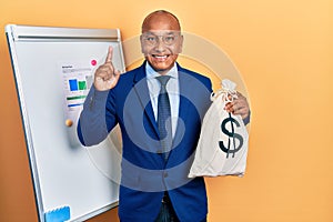 Middle age latin man wearing business suit holding dollars bag surprised with an idea or question pointing finger with happy face,