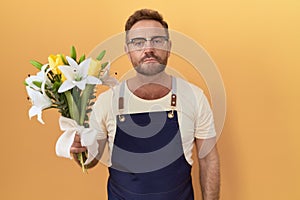 Middle age man with beard florist shop holding flowers relaxed with serious expression on face