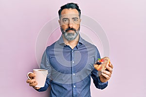 Middle age man with beard and grey hair drinking coffee and eating a pastry relaxed with serious expression on face