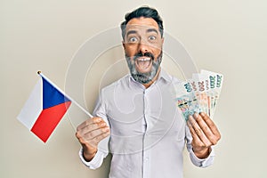 Middle age man with beard and grey hair holding czech republic flag and koruna banknotes celebrating crazy and amazed for success