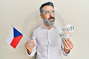 Middle age man with beard and grey hair holding czech republic flag and koruna banknotes smiling looking to the side and staring