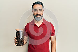 Middle age man with beard and grey hair holding jar with coffee beans looking positive and happy standing and smiling with a