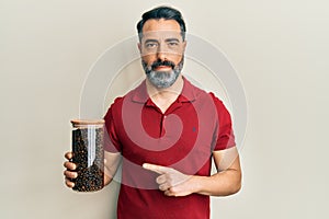 Middle age man with beard and grey hair holding jar with coffee beans smiling happy pointing with hand and finger