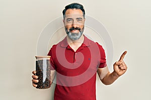 Middle age man with beard and grey hair holding jar with coffee beans smiling happy pointing with hand and finger to the side