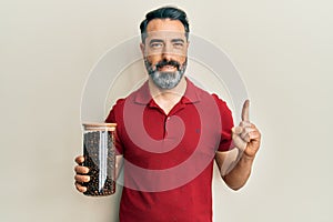 Middle age man with beard and grey hair holding jar with coffee beans smiling with an idea or question pointing finger with happy