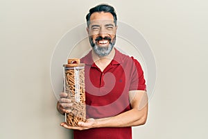 Middle age man with beard and grey hair holding jar with macaroni pasta smiling and laughing hard out loud because funny crazy