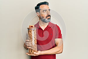 Middle age man with beard and grey hair holding jar with macaroni pasta smiling looking to the side and staring away thinking