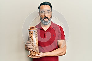 Middle age man with beard and grey hair holding jar with macaroni pasta sticking tongue out happy with funny expression