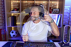 Middle age man with beard playing video games wearing headphones smiling doing phone gesture with hand and fingers like talking on