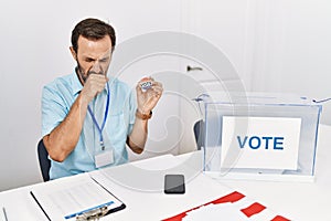 Middle age man with beard sitting by ballot holding i vote badge feeling unwell and coughing as symptom for cold or bronchitis