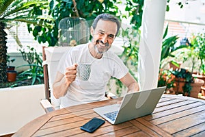 Middle age man with beard smiling happy at the terrace working from home using laptop drinking a cup of coffee