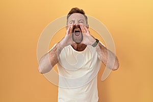 Middle age man with beard standing over yellow background shouting angry out loud with hands over mouth