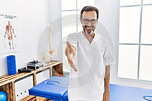 Middle age man with beard working at pain recovery clinic smiling friendly offering handshake as greeting and welcoming