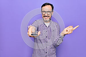 Middle age man changing tv channel using television remote control over purple background smiling happy pointing with hand and