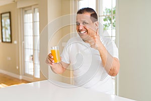 Middle age man drinking a glass of orange juice at home happy with big smile doing ok sign, thumb up with fingers, excellent sign