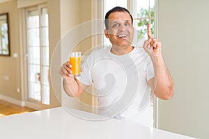Middle age man drinking a glass of orange juice at home surprised with an idea or question pointing finger with happy face, number