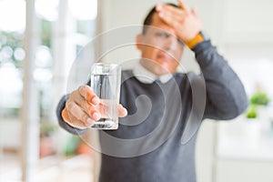 Middle age man drinking glass of water at home stressed with hand on head, shocked with shame and surprise face, angry and