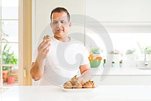 Middle age man eating chocolate chip muffins at home with a confident expression on smart face thinking serious