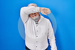 Middle age man with grey hair standing over blue background covering eyes with arm, looking serious and sad