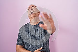 Middle age man with grey hair standing over pink background laughing at you, pointing finger to the camera with hand over body,