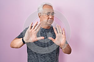 Middle age man with grey hair standing over pink background moving away hands palms showing refusal and denial with afraid and