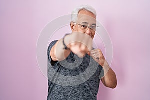 Middle age man with grey hair standing over pink background punching fist to fight, aggressive and angry attack, threat and