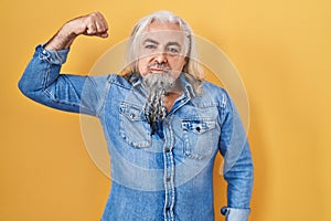 Middle age man with grey hair standing over yellow background strong person showing arm muscle, confident and proud of power