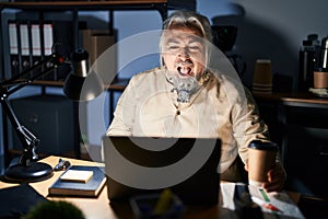 Middle age man with grey hair working at the office at night angry and mad screaming frustrated and furious, shouting with anger