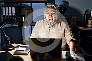 Middle age man with grey hair working at the office at night angry and mad screaming frustrated and furious, shouting with anger