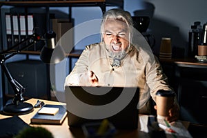 Middle age man with grey hair working at the office at night pointing displeased and frustrated to the camera, angry and furious
