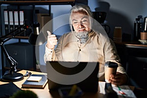 Middle age man with grey hair working at the office at night pointing to the back behind with hand and thumbs up, smiling