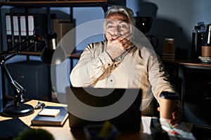 Middle age man with grey hair working at the office at night shocked covering mouth with hands for mistake