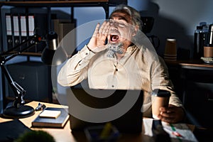 Middle age man with grey hair working at the office at night shouting and screaming loud to side with hand on mouth