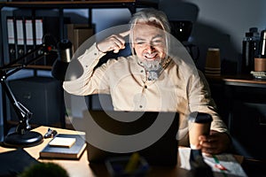 Middle age man with grey hair working at the office at night smiling pointing to head with one finger, great idea or thought, good