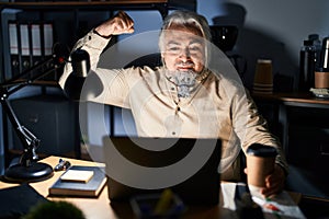 Middle age man with grey hair working at the office at night strong person showing arm muscle, confident and proud of power