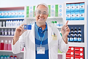 Middle age man with grey hair working at pharmacy drugstore holding credit card smiling happy pointing with hand and finger to the