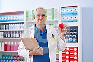 Middle age man with grey hair working at pharmacy drugstore holding red heart relaxed with serious expression on face