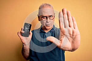 Middle age man holding broken smartphone showing cracked screen over yellow background with open hand doing stop sign with serious