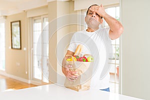 Middle age man holding groceries shopping bag at home stressed with hand on head, shocked with shame and surprise face, angry and