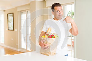 Middle age man holding groceries shopping bag at home with surprise face pointing finger to himself