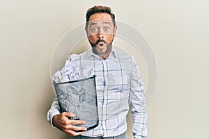 Middle age man holding paper bin full of crumpled papers scared and amazed with open mouth for surprise, disbelief face